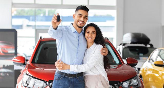 A couple in a car dealership hold up their new keys while standing in front of a new red car.